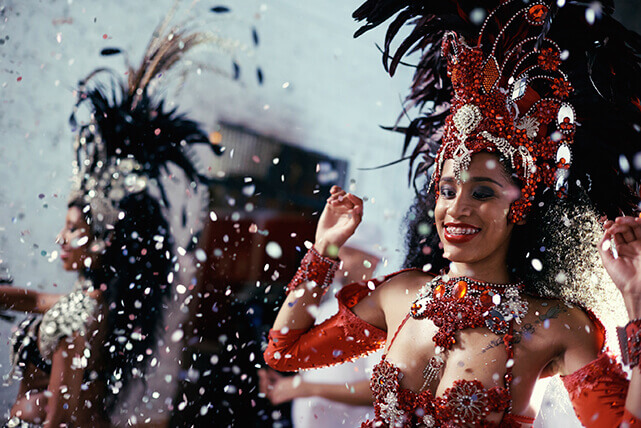 Dançarina sorridente com fantasia vermelha elaborada, cocar de plumas e pedrarias em celebração de carnaval com confete.