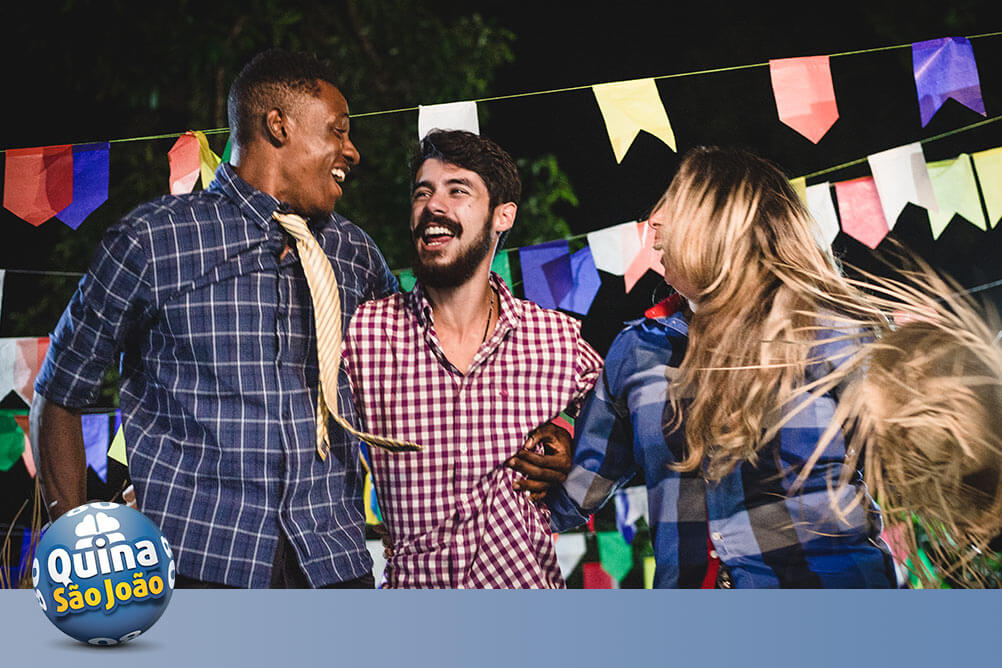 Imagem com três pessoas festeiras vestidas a caráter de São João com bandeirolas juninas ao fundo. Falam sobre os números populares da Quina de São João.