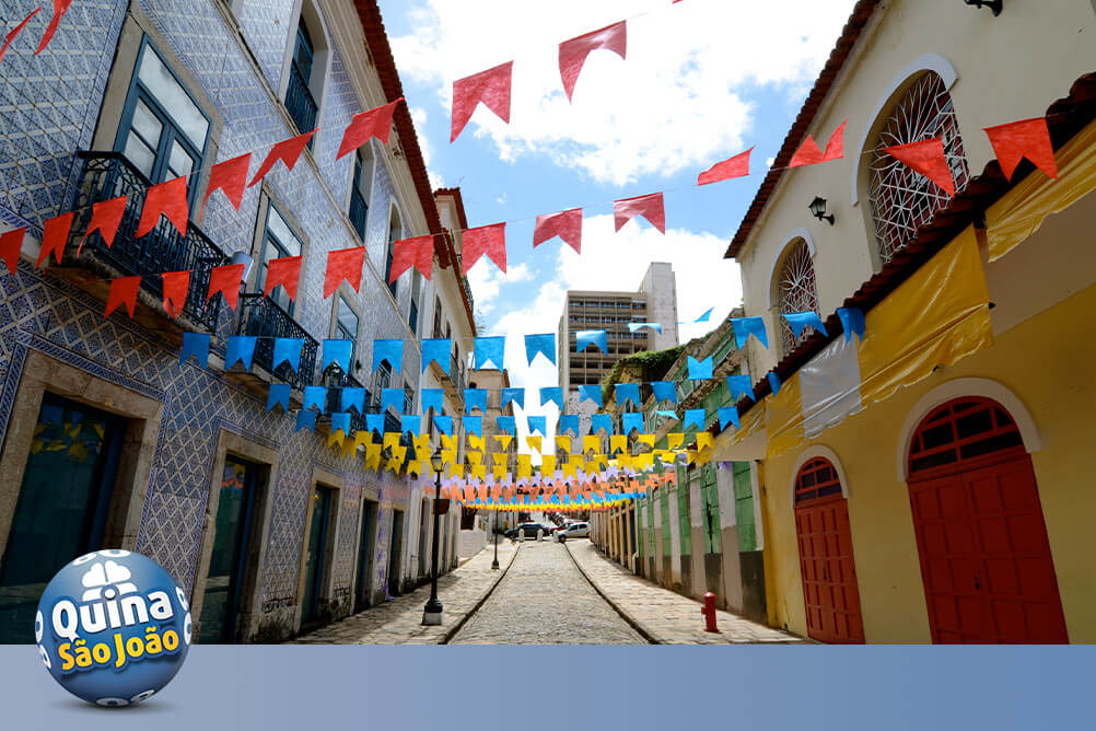 Imagem de uma rua decorada com bandeirolas juninas, casas coloridas e uma casa de apostas que ensina como jogar na Quina de São João.