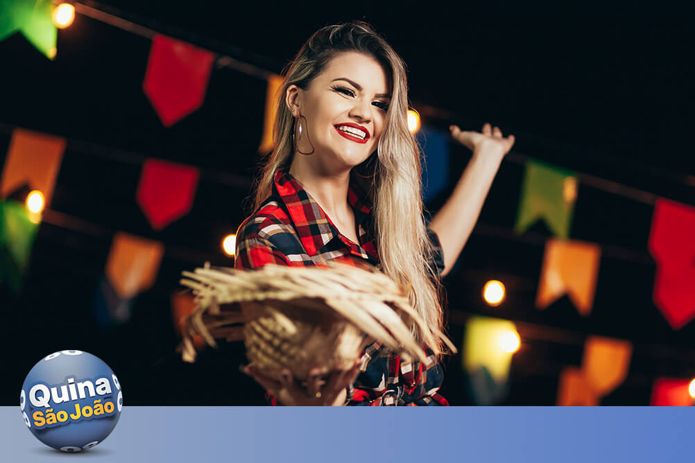 Mulher vestida com roupa xadrez para festa junina, segurando um chapéu de palha com bandeirolas coloridas ao fundo, celebra ser ganhador no sorteio Quina de São João.