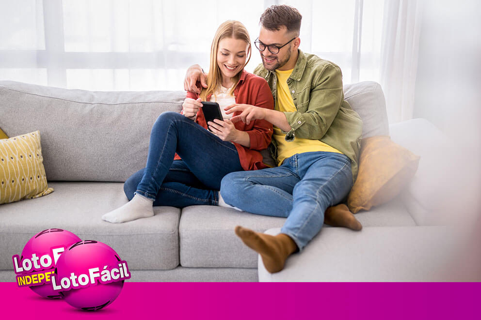 Casal feliz consulta calendário de concursos da Lotofácil pelo celular no sofá de casa.