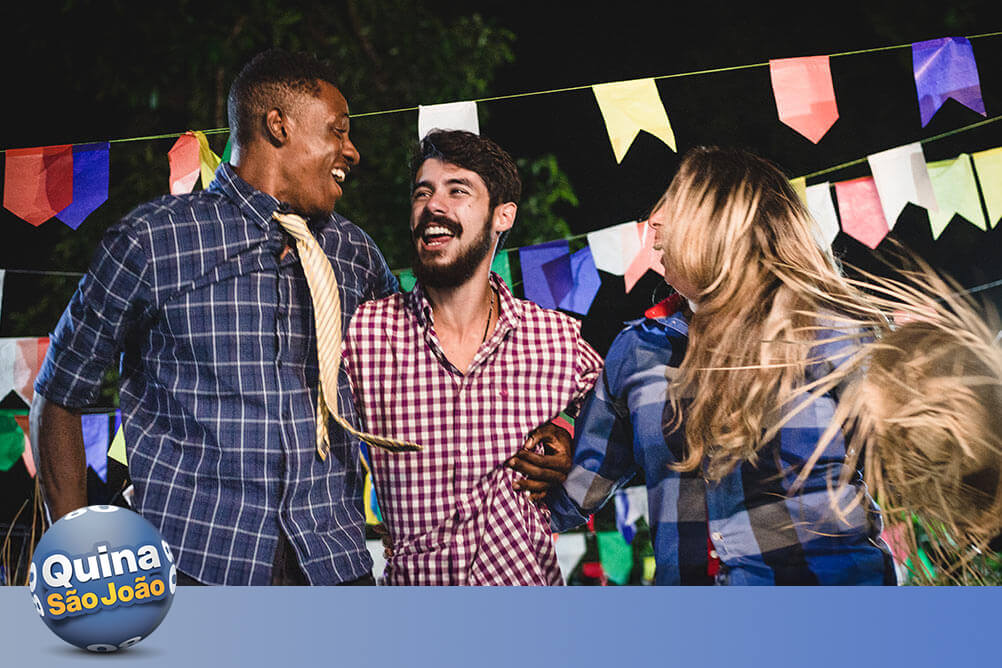 Imagem com três pessoas festeiras vestidas a caráter de São João com bandeirolas juninas ao fundo. Falam sobre os números populares da Quina de São João.